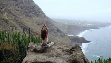 Practicing-yoga-amidst-island-mountains,-a-young-woman-sits-on-a-mountaintop-rock,-meditating-in-Lotus-posture,-with-a-view-of-the-ocean-below